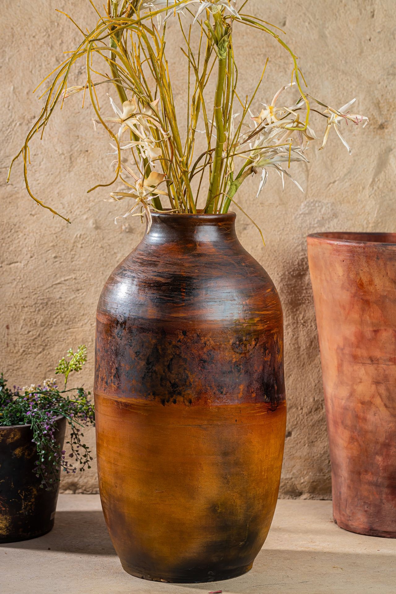 Large ceramic vase with dry yellow and white flowers placed on a rustic background with other clay pots.