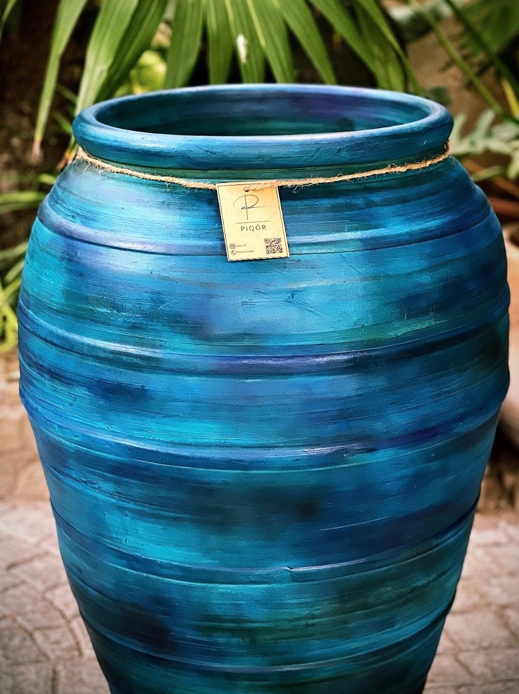 Large blue ceramic pot with a small label attached, placed outdoors with green leaves in the background.