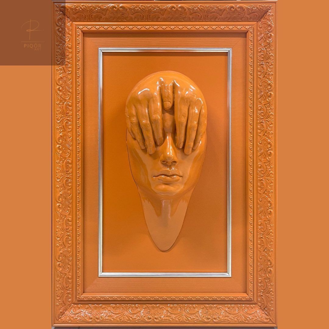 Orange wall art of a face with hands covering eyes inside an ornate frame.