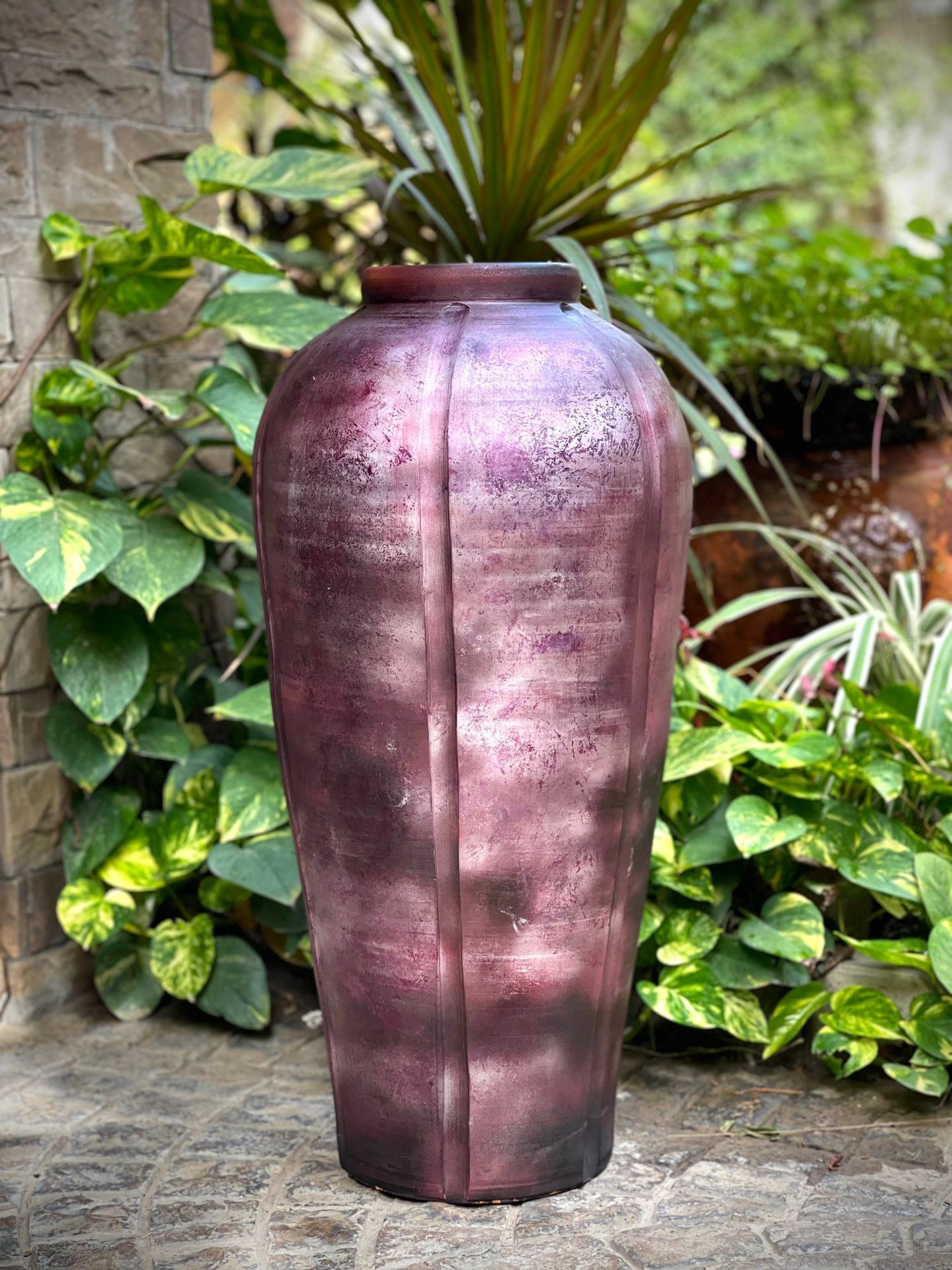 Tall purple vase with a textured surface placed in an outdoor garden setting surrounded by greenery.